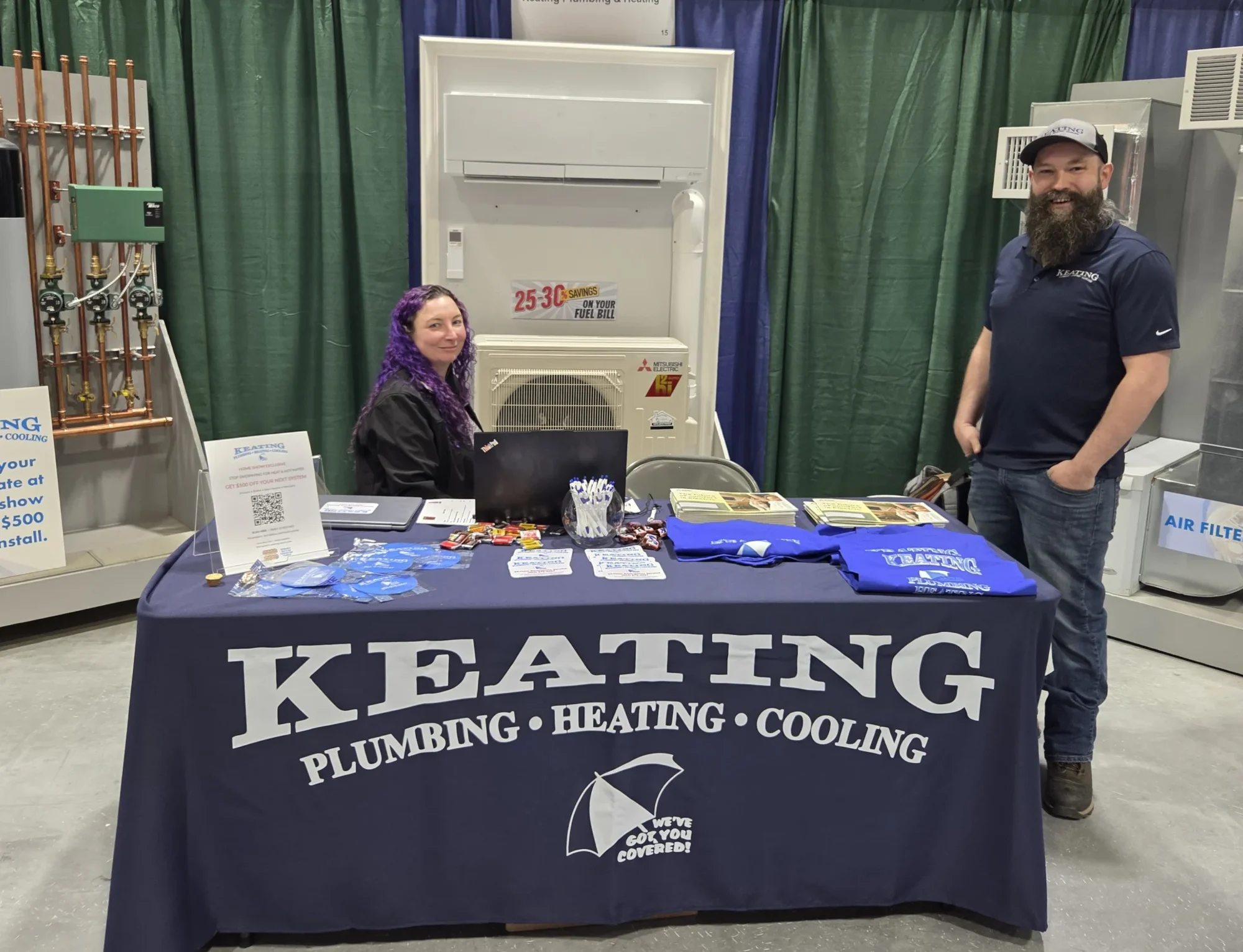 Booth at a tradeshow with a large KEATING banner tablecloth, two people smiling beside plumbing equipment.