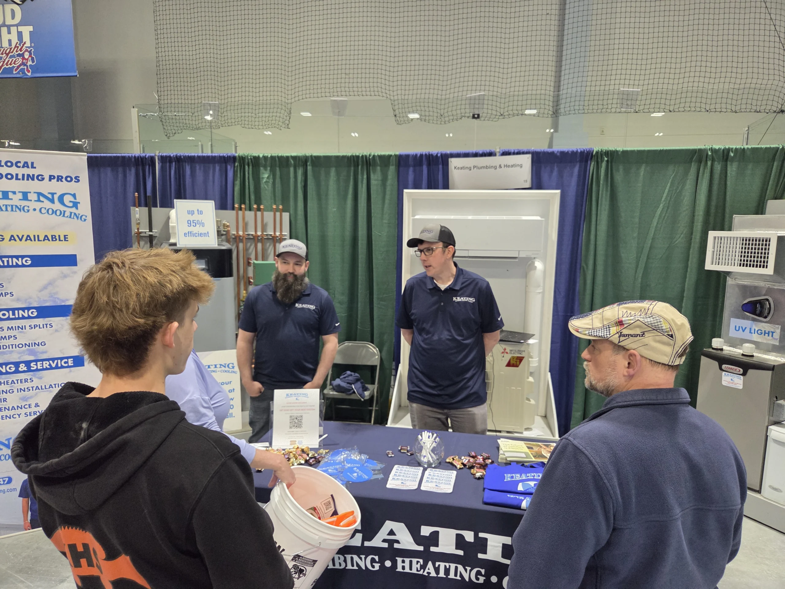 At a trade-show booth, two men in navy shirts stand behind a display with HVAC/heating signs and green curtains, talking to attendees.