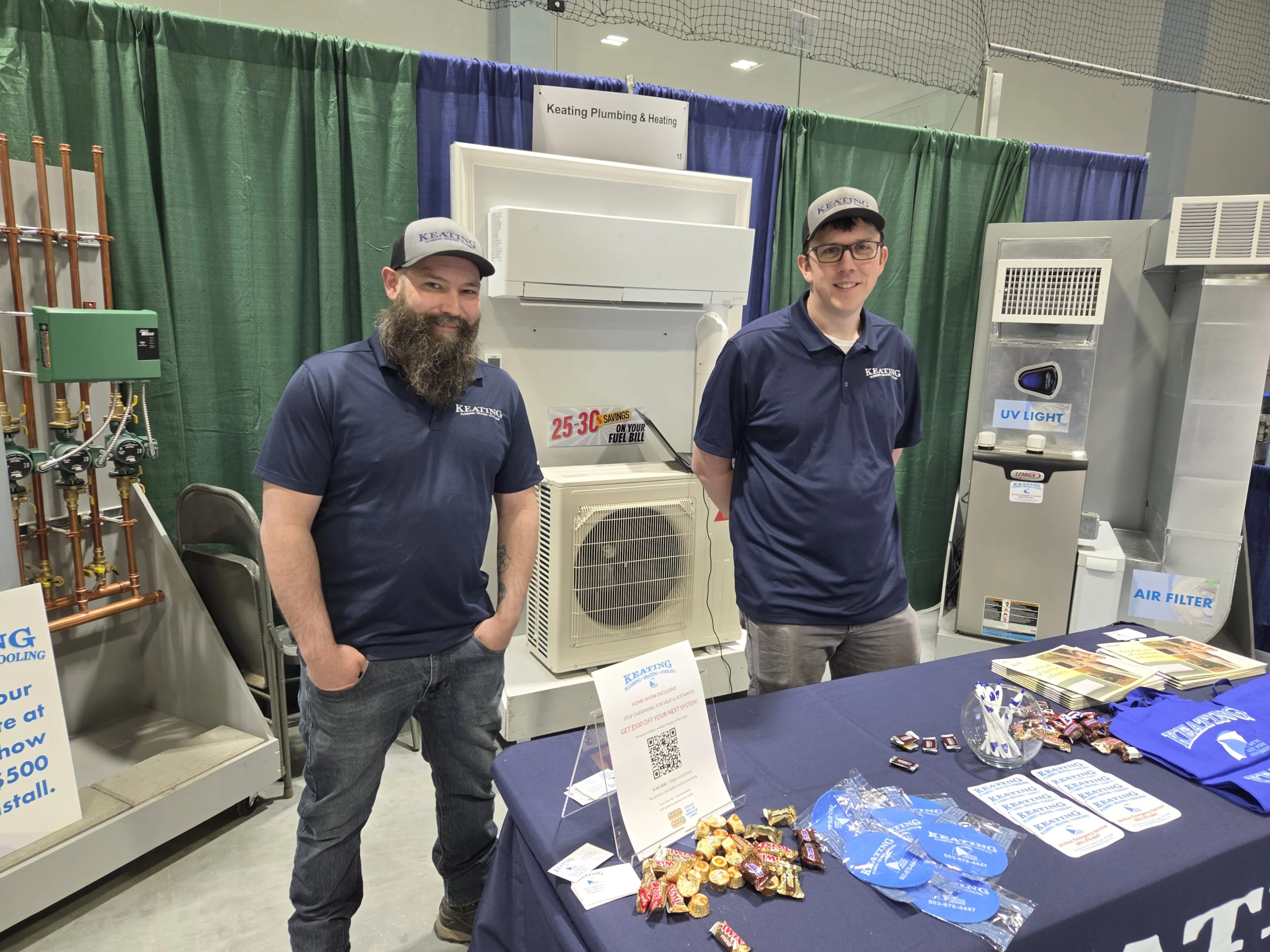 Two men in navy polo shirts and baseball caps stand at an HVAC equipment booth, smiling at the camera with machinery and ducts behind them.
