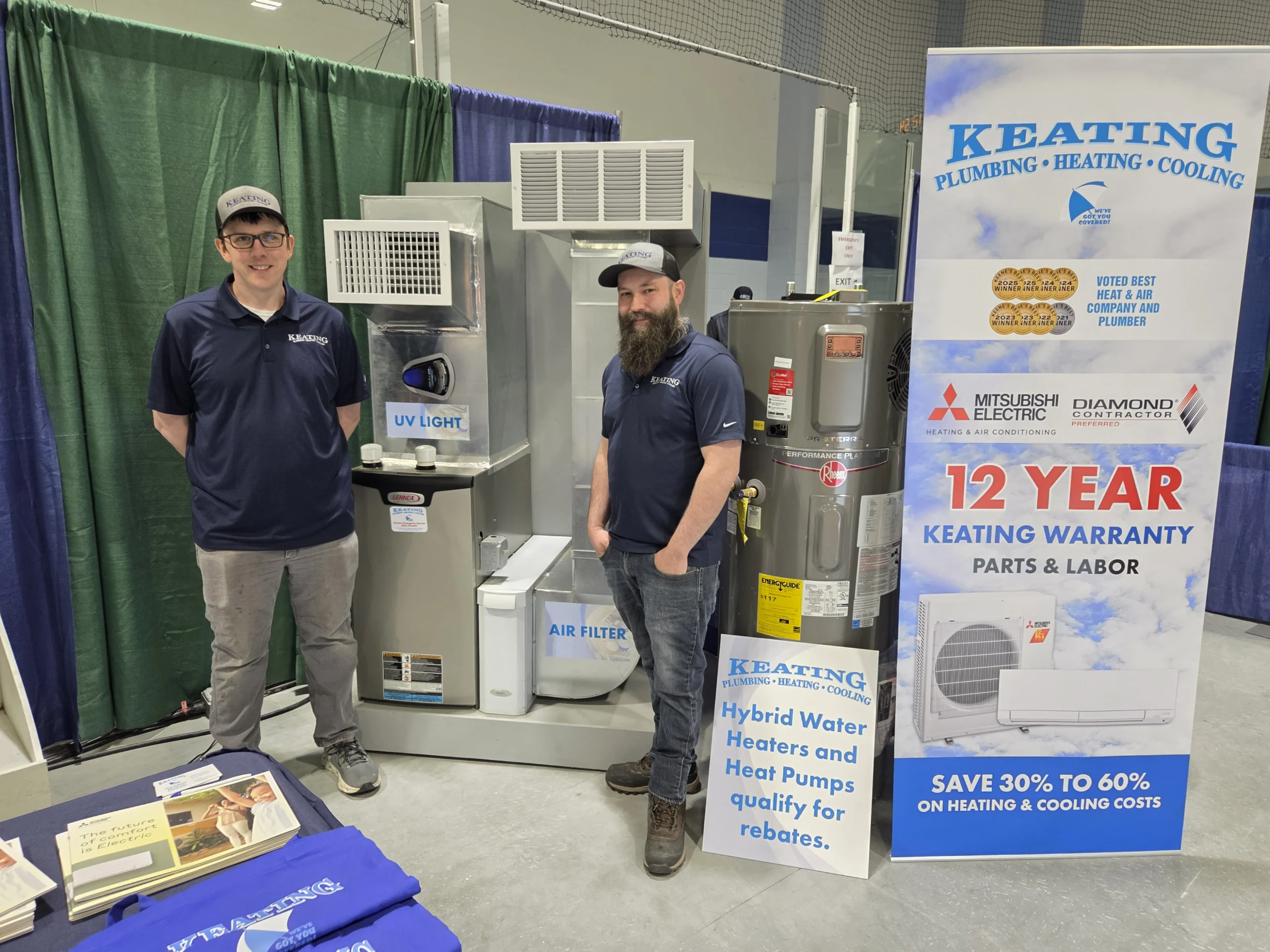 Two Keating technicians stand beside HVAC equipment at a booth, smiling, with a blue-and-white promotional banner nearby.