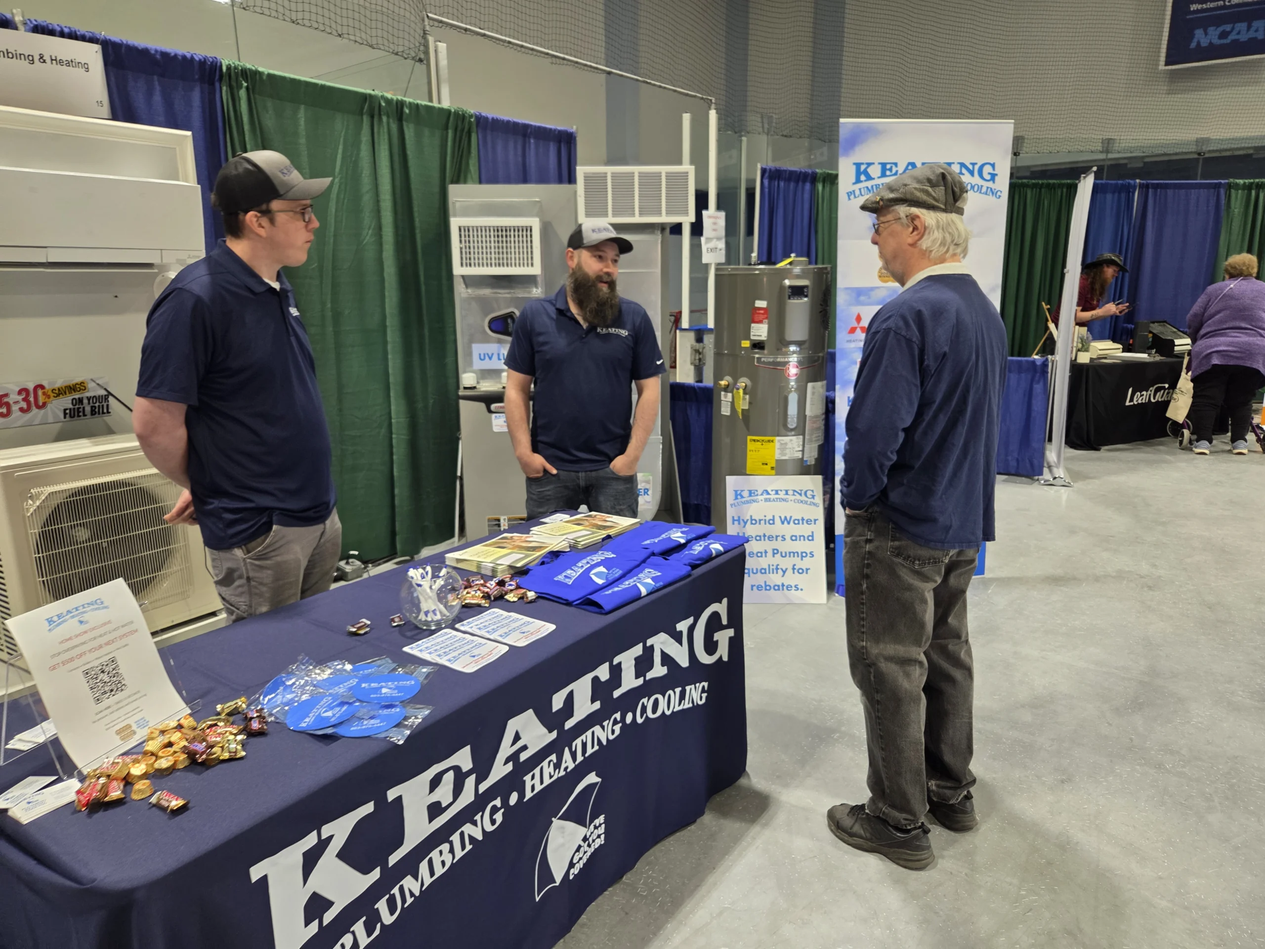Three men in navy shirts stand at a trade show booth with a large blue tablecloth and brochures.